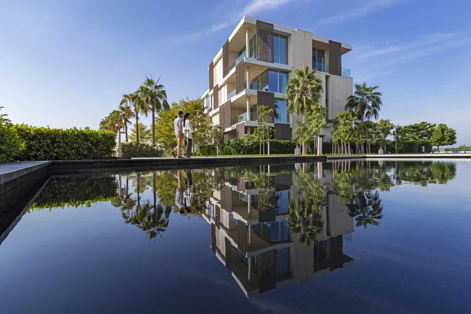 Een modern resort met palmbomen die weerspiegeld worden in een rustige waterpartij; twee mensen lopen over het pad langs het water onder een helderblauwe hemel.